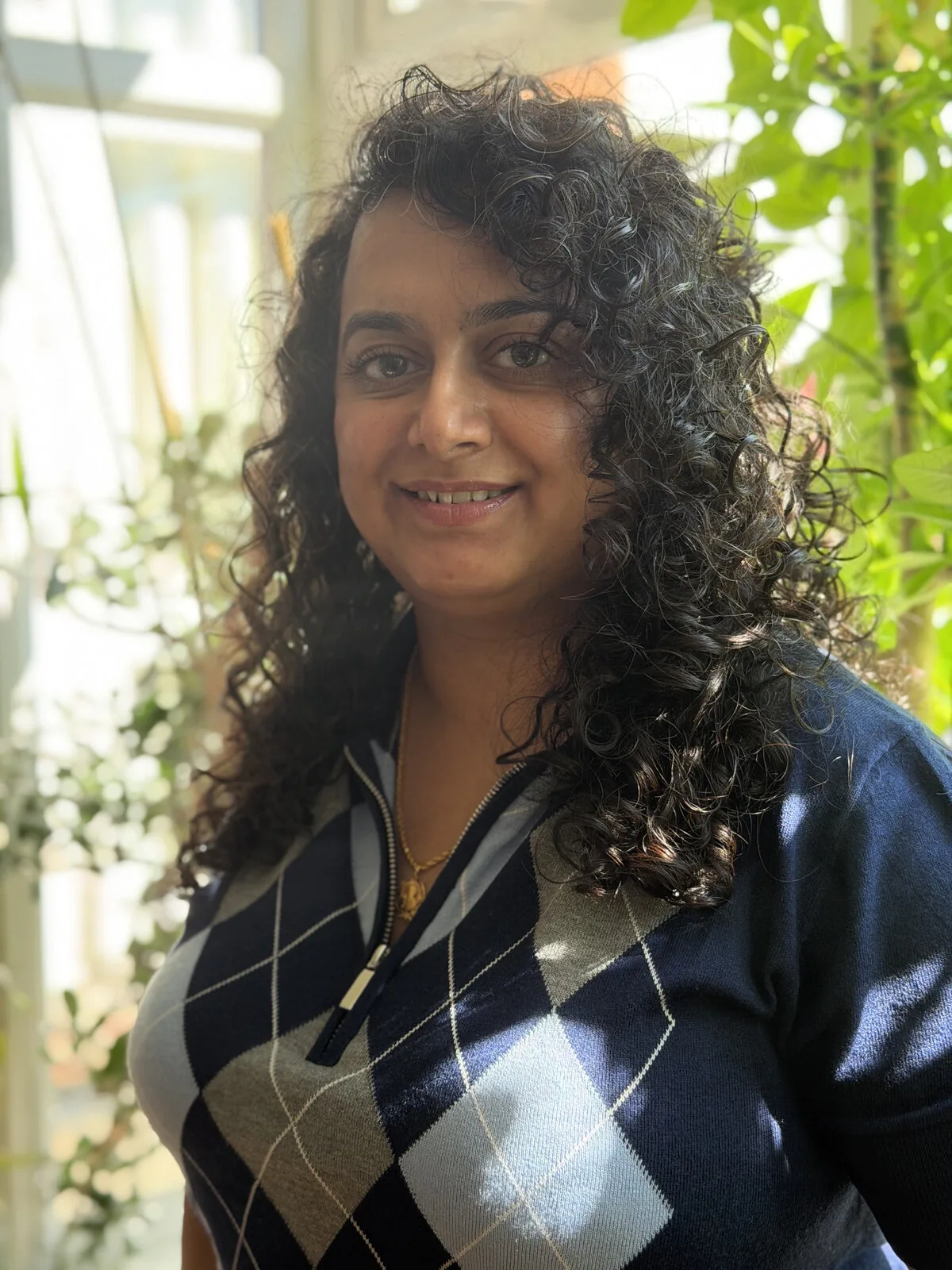 Portrait of Priyaneet Kainth smiling outdoors, wearing a navy argyle-pattern zip-up sweater, with leafy greenery in the background.